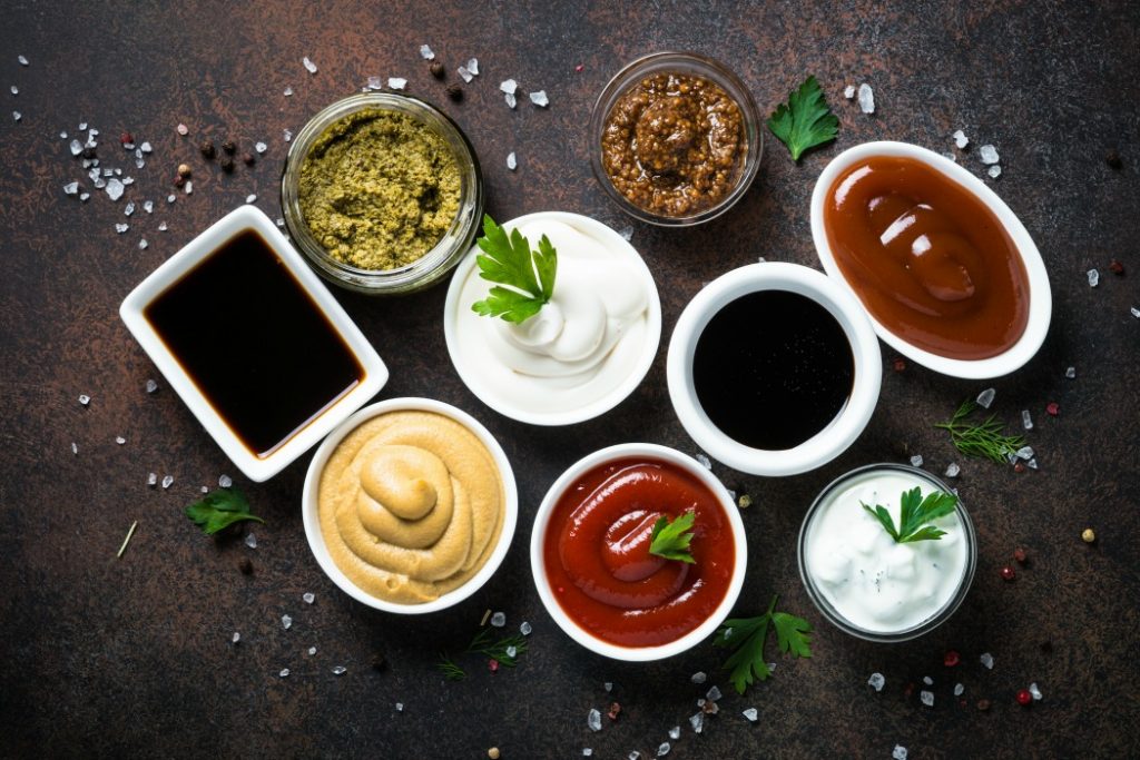 Set of sauces - ketchup, mayonnaise, mustard soy sauce, bbq sauce, pesto, mustard grains and pomegranate sauce on dark rusty stone or metal background. Top view.